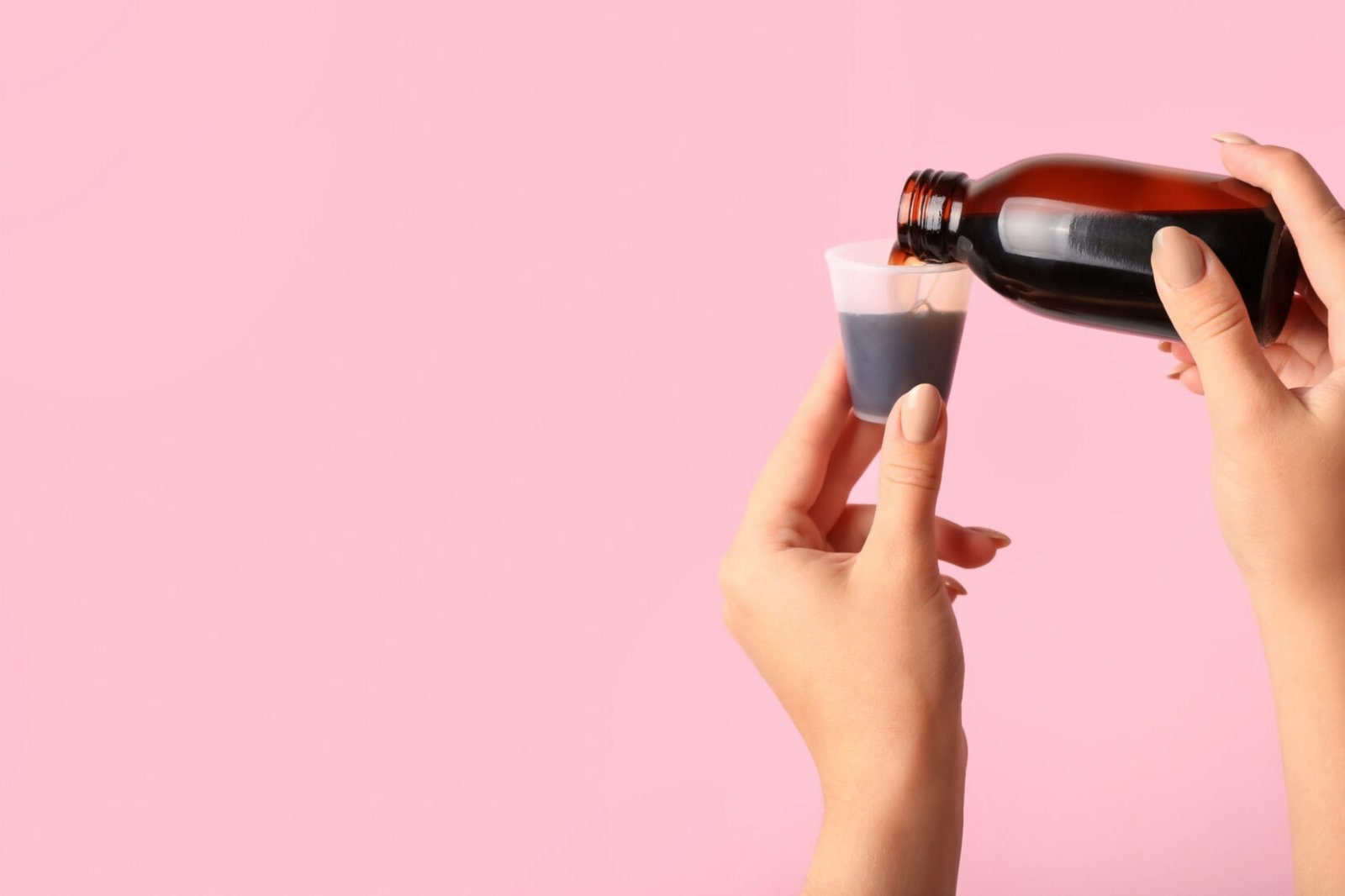 Female hands pouring cough syrup from bottle into cup on pink background with space for text